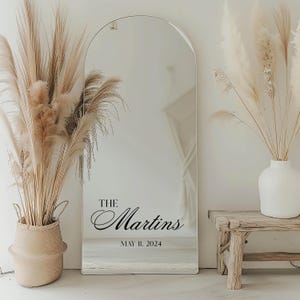 May include: A large, arched mirror with the text "The Martins" and the date "May 11, 2024" in black script. The mirror is surrounded by decorative pampas grass in a woven basket and a white vase, with a small wooden bench.