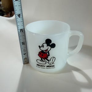 Może przedstawiać: Biały kubek ze szkła mlecznego z grafiką Myszki Miki w czerwonych spodenkach oraz napisem MICKEY MOUSE WALT DISNEY PRODUCTIONS. Miarka umieszczona obok kubka wskazuje wysokość kubka wynoszącą około 8,9 cm.