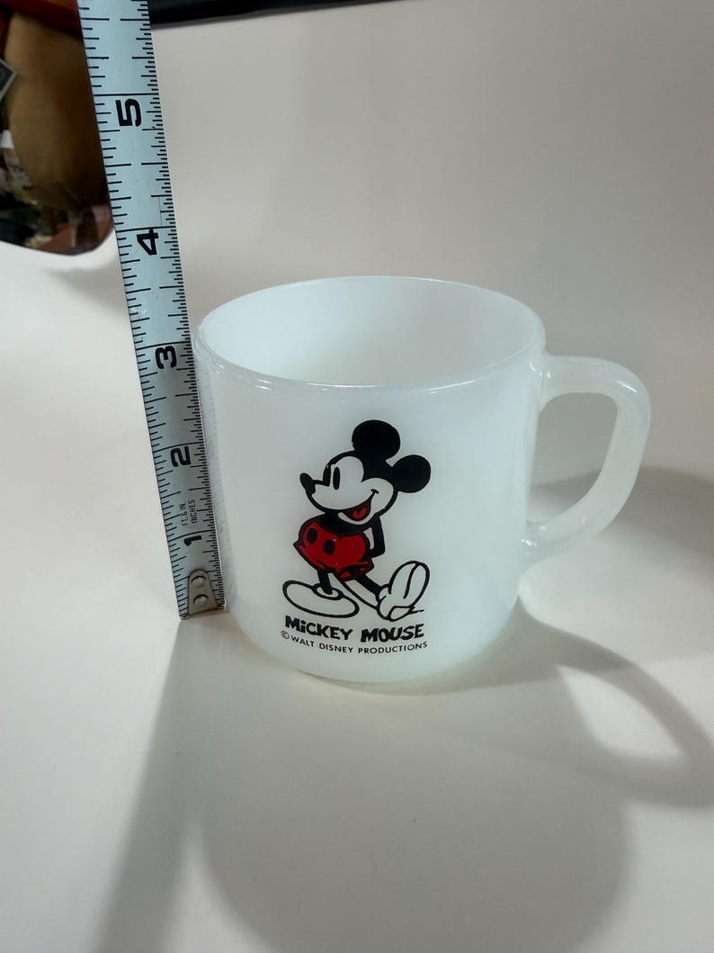 Może przedstawiać: Biały kubek ze szkła mlecznego z grafiką Myszki Miki w czerwonych spodenkach oraz napisem MICKEY MOUSE WALT DISNEY PRODUCTIONS. Miarka umieszczona obok kubka wskazuje wysokość kubka wynoszącą około 8,9 cm.