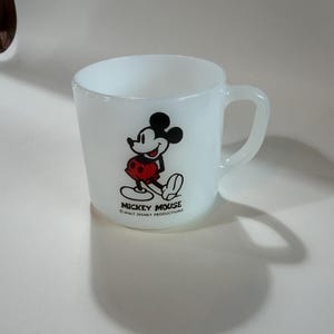 Może przedstawiać: Biały kubek z mlecznego szkła z grafiką Myszki Miki w kolorach czarnym, białym i czerwonym. Napis "MICKEY MOUSE" oraz "WALT DISNEY PRODUCTIONS" jest nadrukowany na czarno pod postacią. Kubek posiada zaokrąglone ucho i gładkie wykończenie.