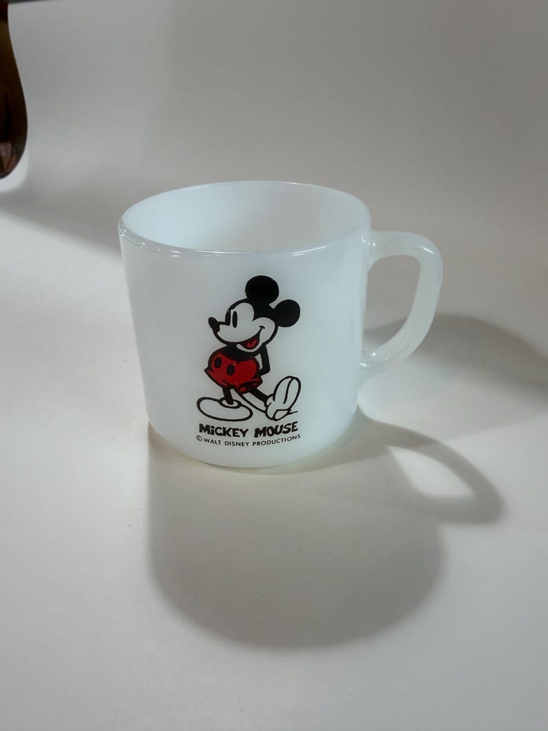 Może przedstawiać: Biały kubek z mlecznego szkła z grafiką Myszki Miki w kolorach czarnym, białym i czerwonym. Napis "MICKEY MOUSE" oraz "WALT DISNEY PRODUCTIONS" jest nadrukowany na czarno pod postacią. Kubek posiada zaokrąglone ucho i gładkie wykończenie.