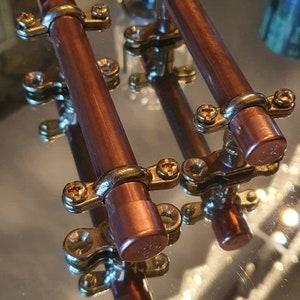 Könnte beinhalten: Zwei Kupfer-Schubladengriffe mit goldenen Akzenten. Die Griffe sind aus Kupferrohr gefertigt und haben eine polierte Oberfläche.