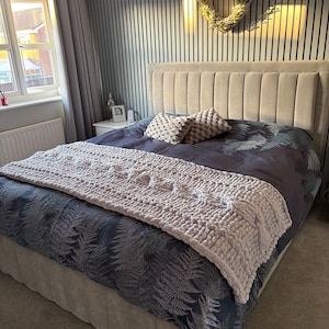 May include: A bedroom with a large bed featuring a gray headboard and a navy blue comforter with a fern pattern. A light gray, chunky knit throw blanket is draped across the bed. Two decorative pillows and a woven wreath complete the decor.