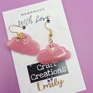 May include: A pair of handmade pink cloud earrings with gold-coloured hooks. The earrings are on a white card with the text "HANDMADE With Love" and a black tag that says "Craft Creations by Emily". The background is purple.