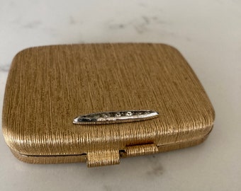 Könnte beinhalten: Ein goldfarbenes Kompakt mit einer strukturierten Oberfläche und einem silbernen Rand mit klaren Steinen.