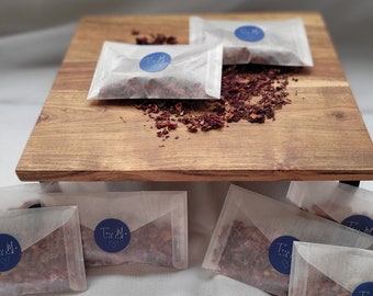 Op de afbeelding: Acht kleine witte papieren zakjes gevuld met gedroogde rozenblaadjes. Elk zakje heeft een blauwe ronde sticker met de tekst "Tea-M. with love". De zakjes zijn gerangschikt op een houten oppervlak.