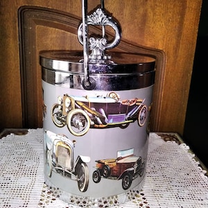 May include: A silver ice bucket with a chrome handle and a vintage car design. The bucket features four different cars, including a blue roadster, a white saloon, a brown roadster, and a red roadster. The bucket is sitting on a white lace tablecloth.