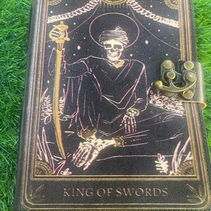 Könnte beinhalten: Schwarzes Lederjournal mit Tarotkarten-Design. Das Cover zeigt eine Skelettfigur mit Stab, die Worte "KING OF SWORDS" und einen dekorativen Verschluss. Das Kunstwerk ist in Gold- und Rosatönen gehalten.