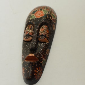 Può includere: Una maschera di legno con sfondo nero e intricati disegni floreali in arancio, verde e oro. La maschera ha un viso scolpito con gli occhi chiusi e una bocca leggermente aperta.