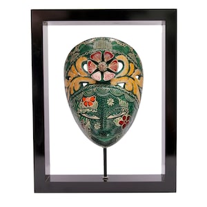Op de afbeelding: Een groen, geel en rood geschilderd houten masker met bloemmotieven wordt getoond in een zwart frame op een standaard.