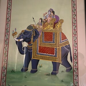 May include: A hand-painted artwork depicting a blue elephant with a red and gold howdah. Four figures are seated in the howdah, including a man with a blue turban and a woman with a red sari. The artwork is framed and has a green background with floral details.
