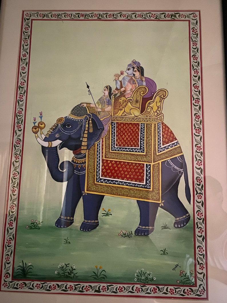 May include: A hand-painted artwork depicting a blue elephant with a red and gold howdah. Four figures are seated in the howdah, including a man with a blue turban and a woman with a red sari. The artwork is framed and has a green background with floral details.