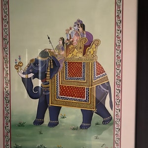 May include: A traditional Indian painting depicting a blue elephant carrying a decorated howdah with four figures inside. The elephant is adorned with gold and red embellishments, and the howdah is decorated with intricate patterns. The painting is set against a light green background with a floral border.