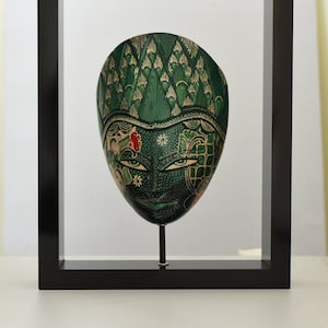 Op de afbeelding: Een groen en gouden geschilderd houten masker met ingewikkelde details, tentoongesteld in een zwart frame.