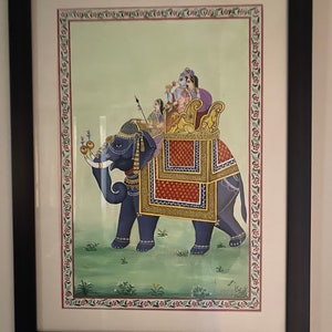 May include: A framed painting depicting a blue elephant carrying a golden howdah with four people riding on top. The elephant is standing on a green field with flowers. The painting is surrounded by a decorative border.