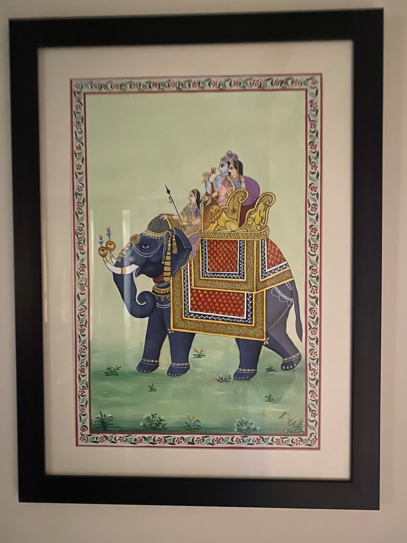 May include: A framed painting depicting a blue elephant carrying a golden howdah with four people riding on top. The elephant is standing on a green field with flowers. The painting is surrounded by a decorative border.