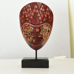 Op de afbeelding: Een houten masker met ingewikkelde handgeschilderde details in rood, groen en goud. Het masker toont een gestileerd menselijk gezicht met bloemmotieven en is gemonteerd op een zwarte houten standaard.