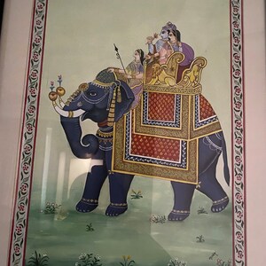 May include: A traditional Indian painting depicting a blue elephant carrying a howdah with four people inside. The elephant is adorned with gold and red decorations. The howdah is decorated with red, gold, and blue patterns. The painting is framed in a white frame with a decorative border.