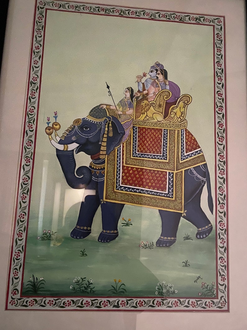 May include: A traditional Indian painting depicting a blue elephant carrying a howdah with four people inside. The elephant is adorned with gold and red decorations. The howdah is decorated with red, gold, and blue patterns. The painting is framed in a white frame with a decorative border.