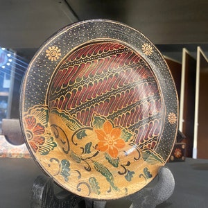 Könnte beinhalten: Ein dekorativer Holzteller mit einem schwarzen, roten, orangen und grünen Batik-Muster. Der Teller steht auf einem schwarzen Holzständer.