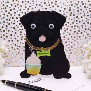 May include: A black pug dog-shaped birthday card with googly eyes, a gold collar, and a cupcake. The card has a green tag that says "happy birthday". A card in the background says "IT'S YOUR BIRTHDAY!"