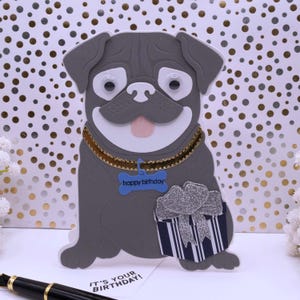 May include: A gray pug holding a gift box with a silver bow. The pug has googly eyes and a blue collar with a bone tag that says "happy birthday".