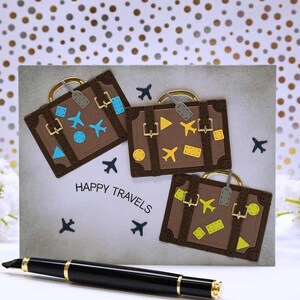 May include: A handmade greeting card with three brown suitcase designs, each adorned with travel-themed embellishments. The card features the text "HAPPY TRAVELS" and is accompanied by a black pen with gold accents. The background has a dotted pattern.