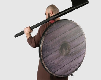 May include: A person wearing a brown tunic and grey trousers is holding a wooden shield and a black axe over their shoulder.