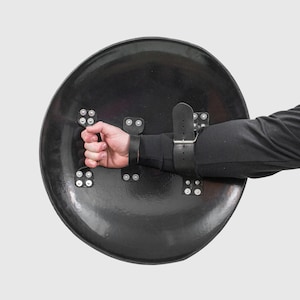 May include: A black round shield with a leather strap and metal rivets. The shield is held by a hand wearing a black glove.