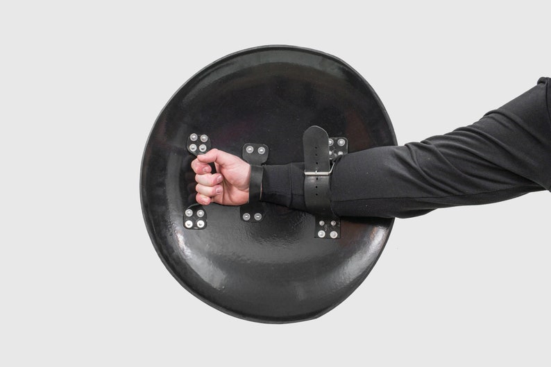May include: A black round shield with a leather strap and metal rivets. The shield is held by a hand wearing a black glove.