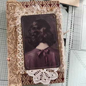 Può includere: Un diario fatto a mano con una copertina dal motivo geometrico rosso e marrone chiaro, decorato con strati di pizzo beige, tessuto sfilacciato e un centrino all'uncinetto bianco. Una stampa rettangolare mostra la schiena di una persona bruna.