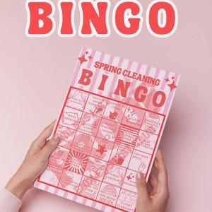 Könnte beinhalten: Eine rosa und weiße "Frühjahrsputz Bingo" Spielkarte mit rotem Titel. Die Karte zeigt Reinigungsaufgaben wie "Fenster putzen" und "Einen Raum entrümpeln". Die Karte wird von Händen vor einem rosa Hintergrund gehalten.
