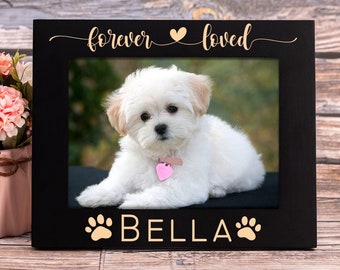 May include: Black picture frame with a white dog inside. The frame has the text "forever loved" with a heart symbol and "BELLA" with paw prints on either side.