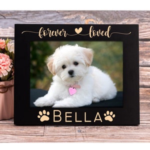 May include: Black picture frame with a white dog inside. The frame has the text "forever loved" with a heart symbol and "BELLA" with paw prints on either side.