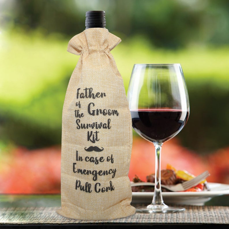 May include: A burlap wine bottle bag with the text "Father of the Groom Survival Kit" and a mustache graphic. A glass of red wine sits next to the bottle, with a blurred outdoor background.