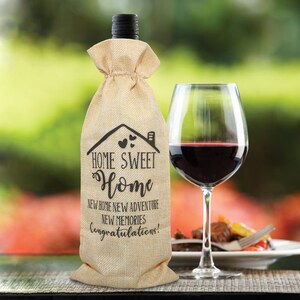 May include: A burlap wine bottle bag with the text "Home Sweet Home" and "New Home New Adventure New Memories Congratulations!" printed on it. The bag is sitting on a table with a glass of red wine and a plate of food.