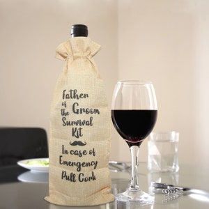 May include: A wine bottle in a burlap bag with the text "Father of the Groom Survival Kit" and a mustache graphic. A glass of red wine and a wine opener are next to the bottle.
