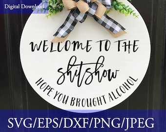 May include: White round wooden sign with black text that reads "Welcome to the shitshow Hope you brought alcohol". The sign is decorated with a black and white checkered ribbon and a brown bow.