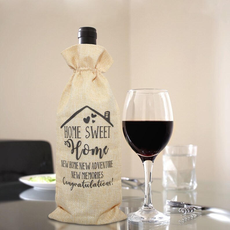 May include: A burlap wine bottle bag with the text "Home Sweet Home" and "New Home New Adventure New Memories Congratulations!" printed on it. The bag is filled with a bottle of wine and is sitting on a table next to a glass of red wine.