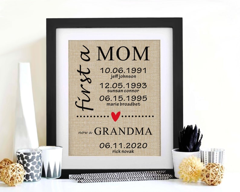 May include: Framed print with the words "first a MOM now a GRANDMA" in black script. The print includes dates and names, with a red heart. The background has a burlap texture, and the frame is black.