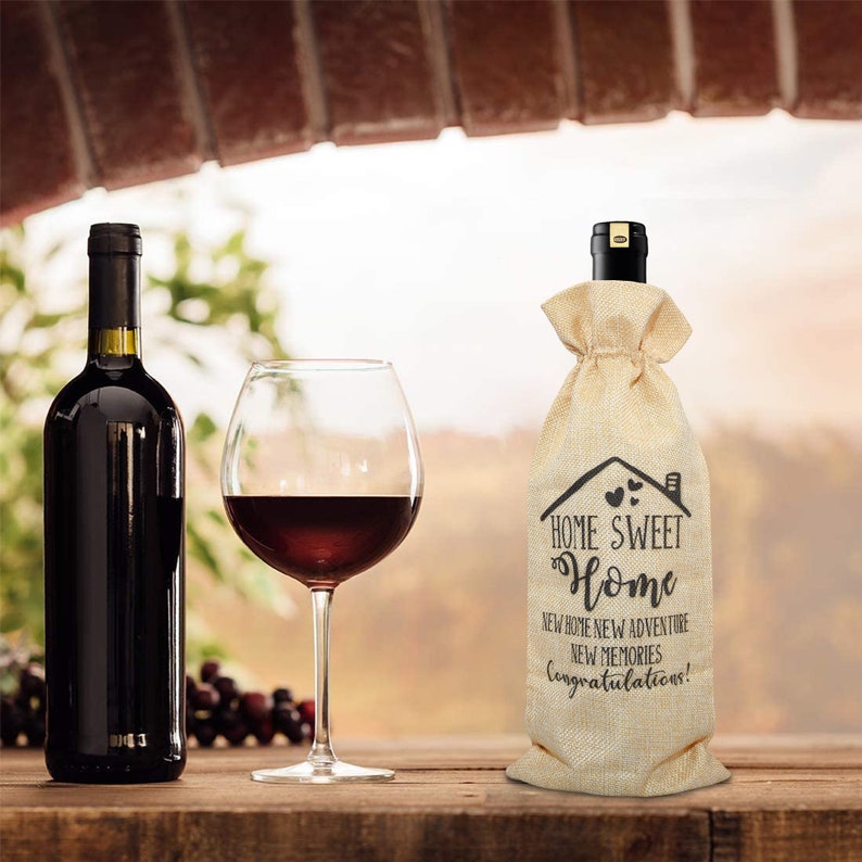 May include: A burlap wine bottle bag with the text "Home Sweet Home" and "New Home New Adventure New Memories! Congratulations!" printed on it. The bag is sitting on a wooden table with a glass of red wine and a bottle of red wine.