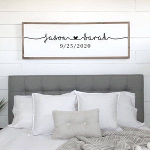 May include: A wooden sign with the text "Jason ♥ Sarah 9/25/2020" in black cursive font. The sign is hanging on a white wall above a grey headboard with white pillows.