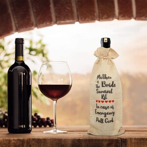 May include: A bottle of red wine, a wine glass with red wine, and a burlap wine bag with the text "Mother of the Bride Survival Kit" and "In case of Emergency Pull Cork" printed on it. The bag has red heart graphics.