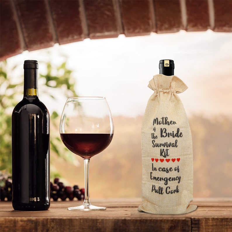 May include: A bottle of red wine, a wine glass with red wine, and a burlap wine bag with the text "Mother of the Bride Survival Kit" and "In case of Emergency Pull Cork" printed on it. The bag has red heart graphics.