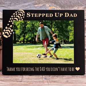 May include: Black wood picture frame with a gold footprint design and the text "Stepped Up Dad" and "Thank you for being the dad you didn't have to be". The frame has a photo of a man and a boy playing soccer.