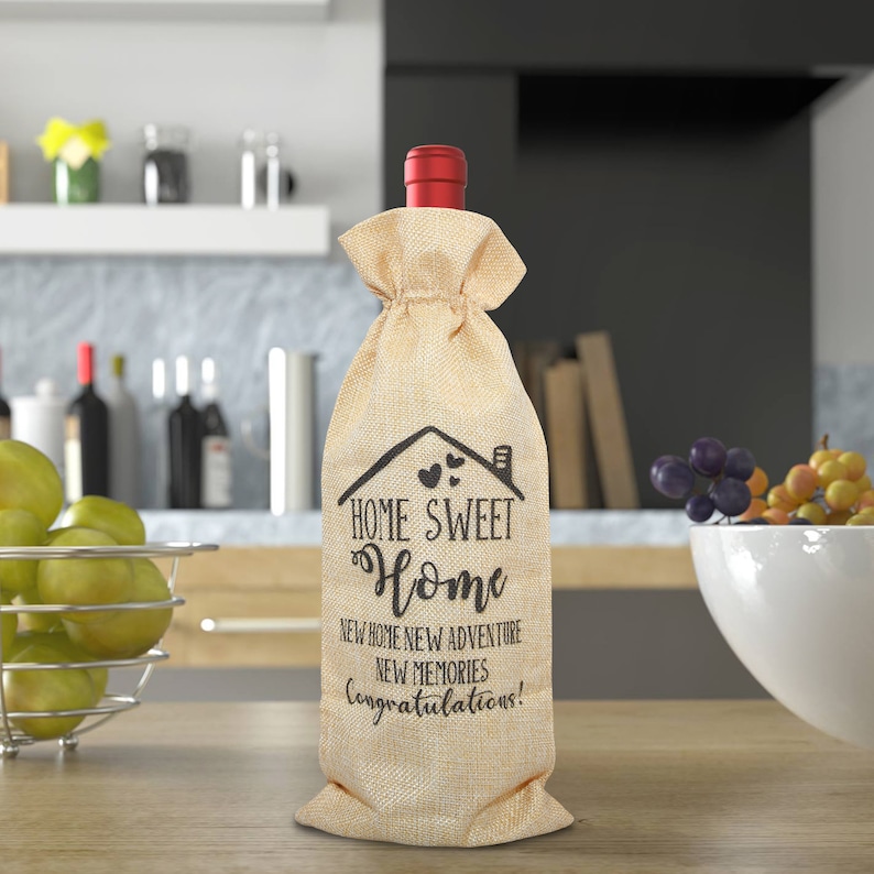 May include: A beige burlap wine bottle bag with black text that reads "Home Sweet Home New Home New Adventure New Memories Congratulations!"