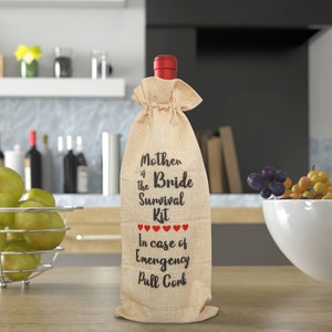 May include: A wine bottle with a red top is encased in a burlap bag. The bag is printed with the text "Mother of the Bride Survival Kit" and "In case of Emergency Pull Cork" in black. Red hearts are printed below the words "Survival Kit".