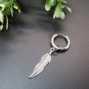 May include: A silver hoop earring with a silver feather charm hanging from it. The earring is on a dark gray background.
