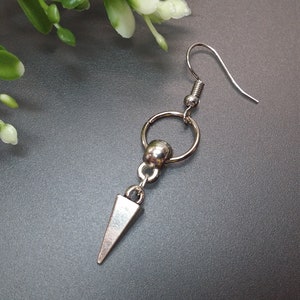 May include: A silver-toned earring with a hook, a small ring, a bead, and a downward-pointing triangular charm. The earring is set against a gray background with a sprig of greenery in the upper left corner.