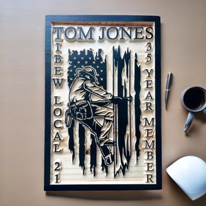 May include: A wooden sign with a black frame and a distressed American flag design. The sign reads "TOM JONES IBEW LOCAL 21 35 YEAR MEMBER". A silhouette of a lineman climbing a pole is in the center of the sign.